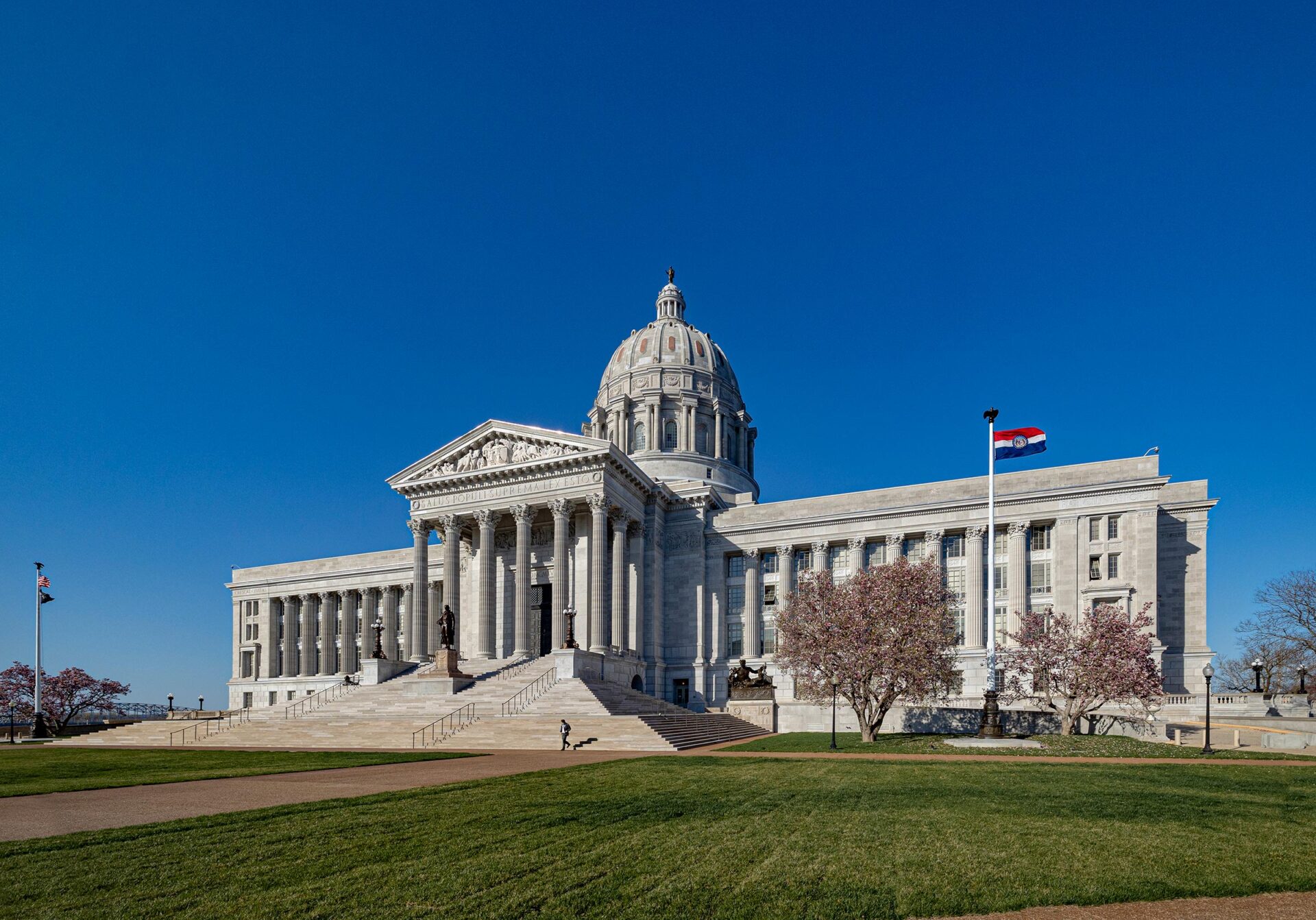 The Missouri State Capitol building in Jefferson City, Missouri under a bright blue sky with cherry blossoms and the Missouri state flag flying